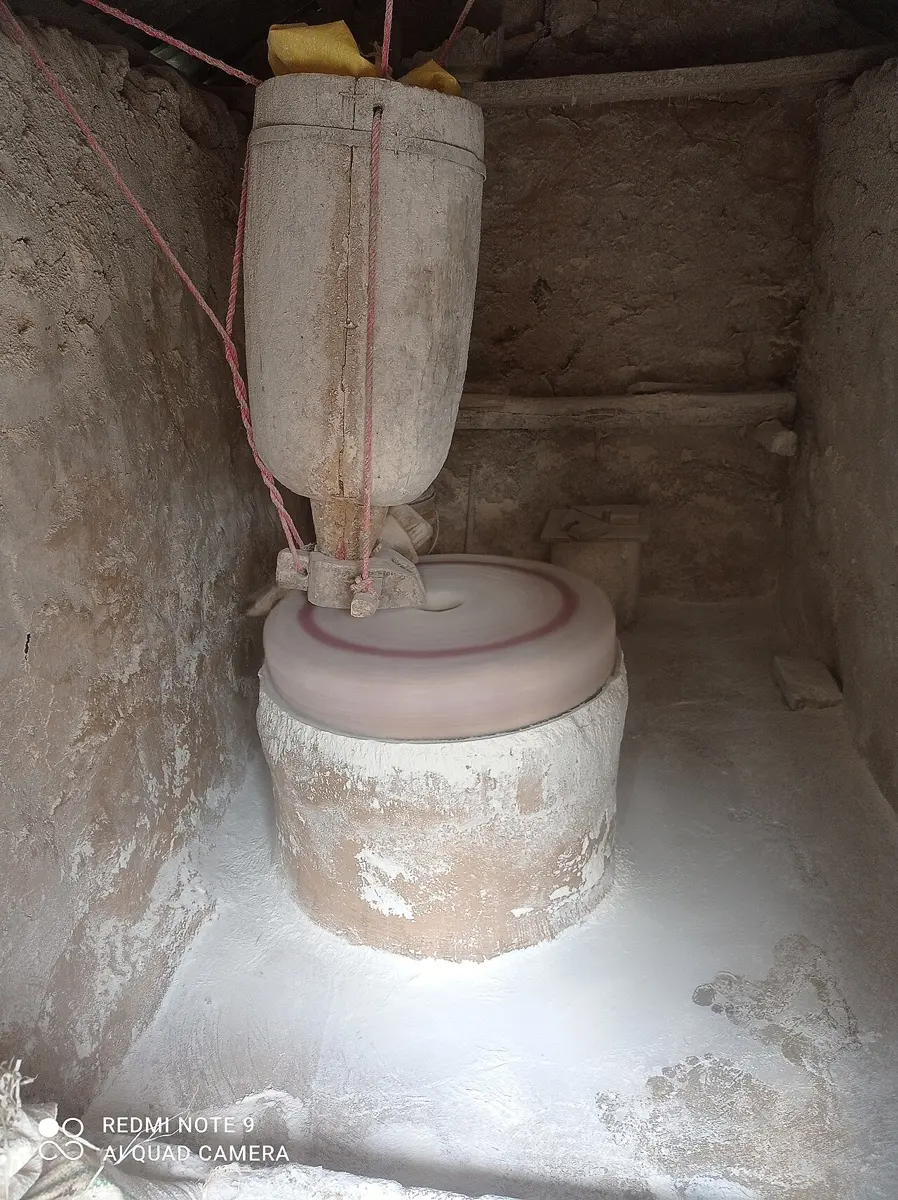 Inside a small mill: grain pours from a wooden hopper onto a round millstone, with white flour around it