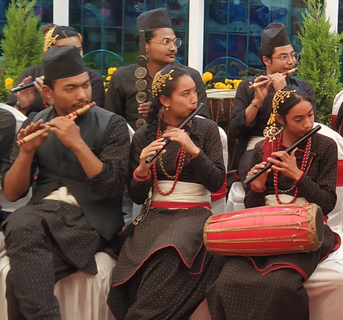 नेपाली पोसाक लगाएर लहरै बसेका युवा कलाकारहरू बाँसको बाँसुरी बजाउँदै; एक जनाको काखमा मादल