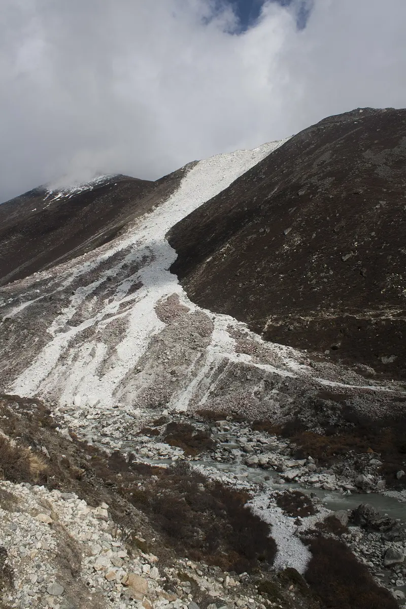 अँध्यारो पहाडको भिरबाट तलतिर झरेको फिक्का फुटेका चट्टानको लामो पट्टी, तल खोलाको छेउमा पङ्खाजस्तो फैलिएको