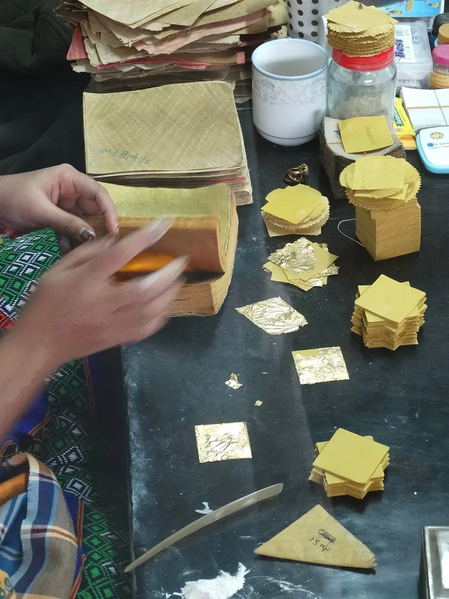 Crinkled, shiny squares of gold leaf and stacks of paper packets on a workshop table, with a worker’s hands.