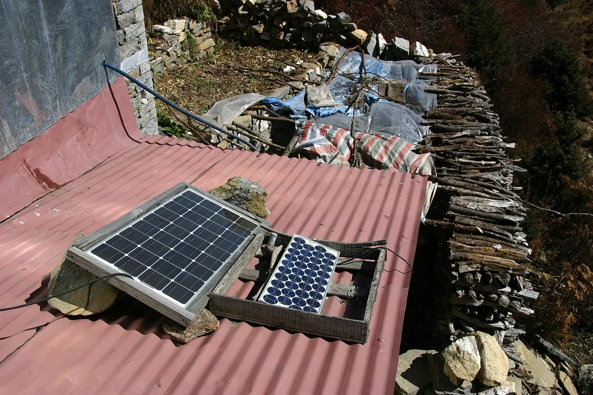 Two small solar panels in wooden frames on the red tin roof of a house in the mountains of Nepal, with a wire running from them, and stacked firewood beside the roof