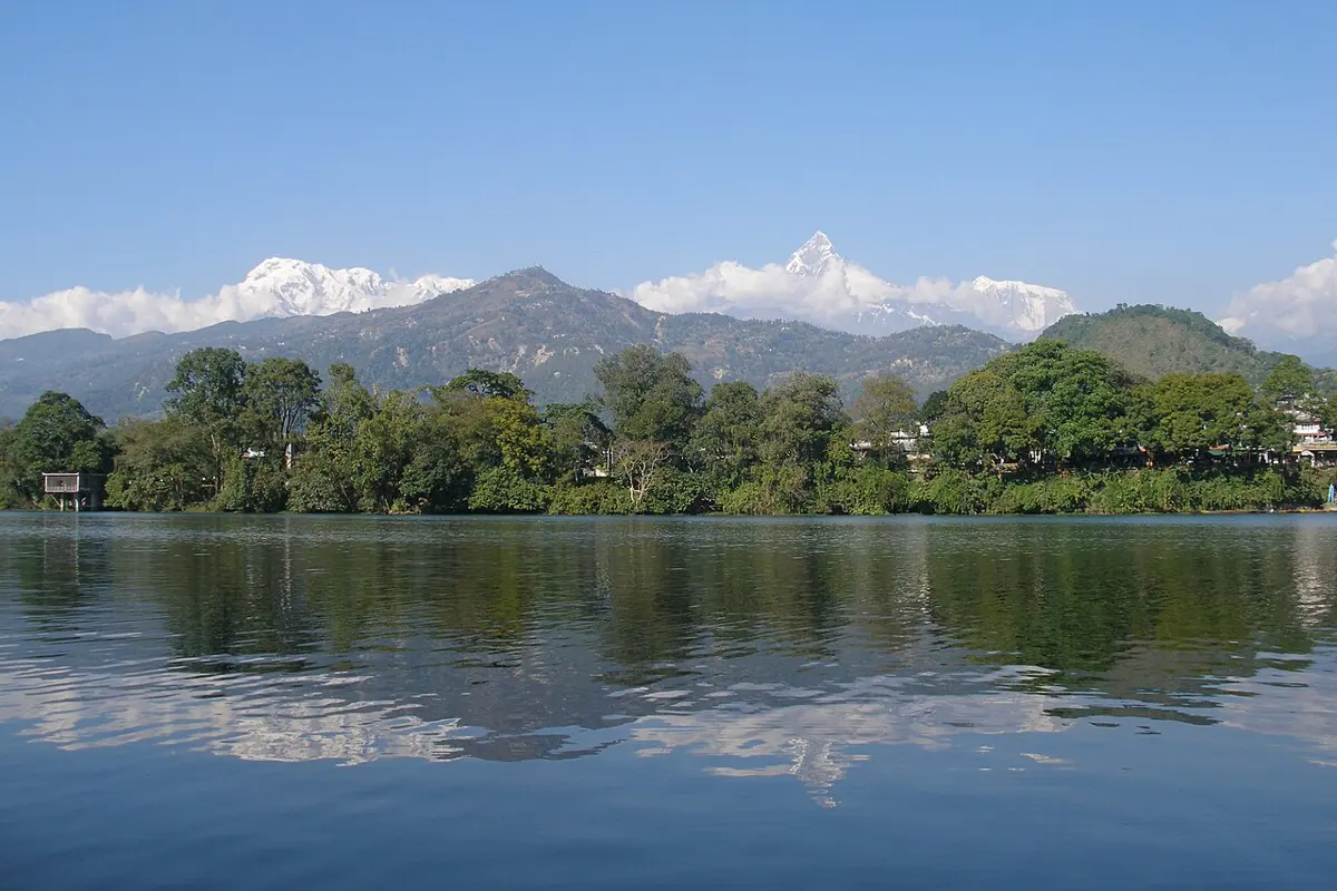 A wide lake with calm blue water, forested hills and snowy mountains behind