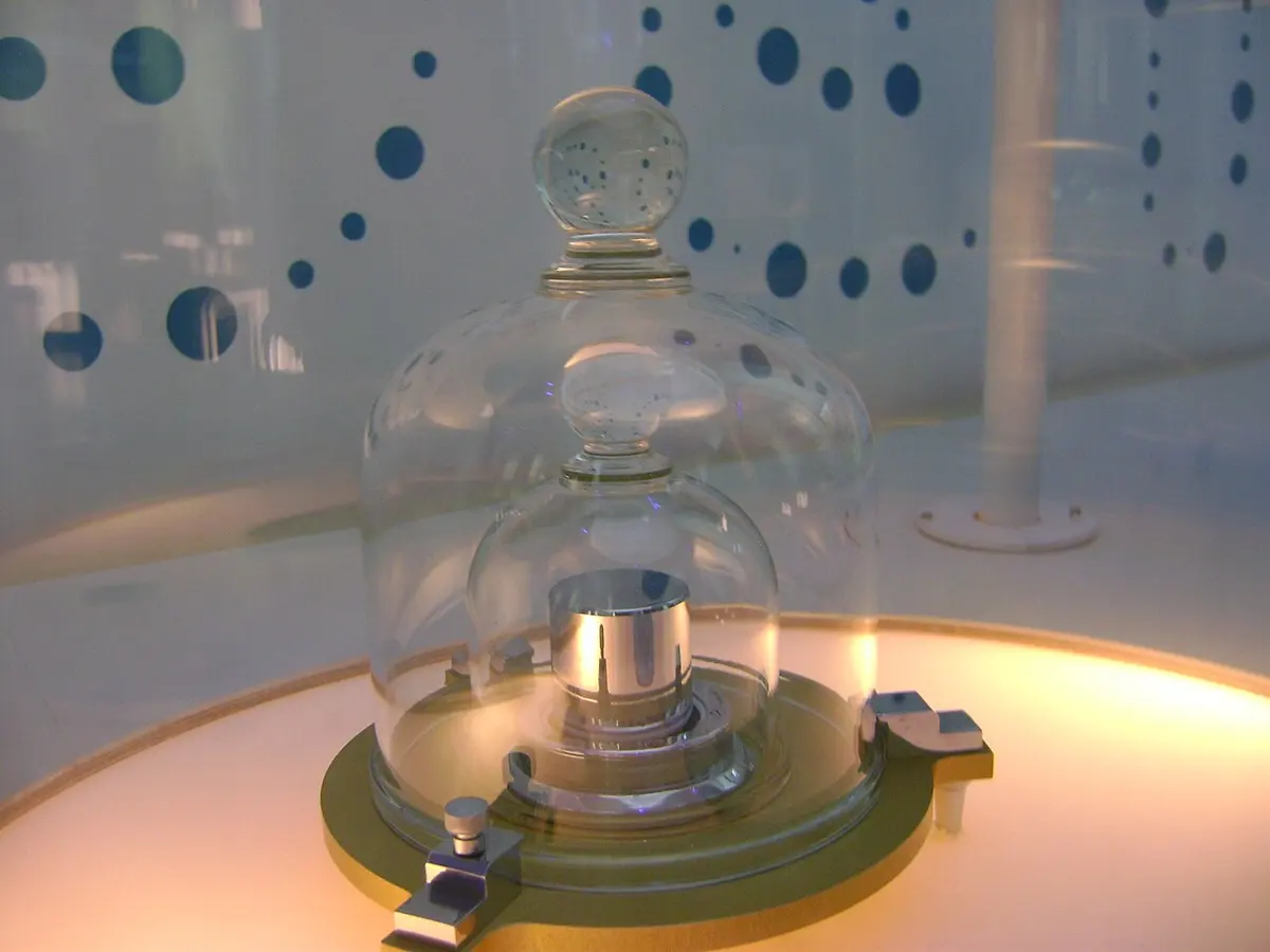 A small shiny metal cylinder under two glass bell jars, on a lit museum stand