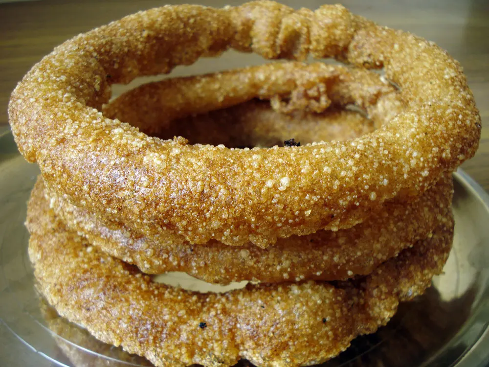 Rings of golden-brown sel roti, sprinkled with sugar, stacked on a plate.