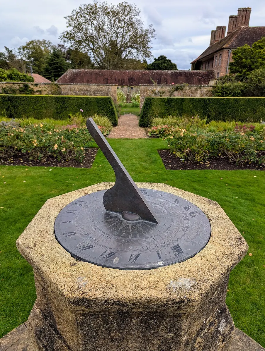 A round metal sundial on a stone pillar in a garden, with a slanting metal blade casting a shadow across the hour marks