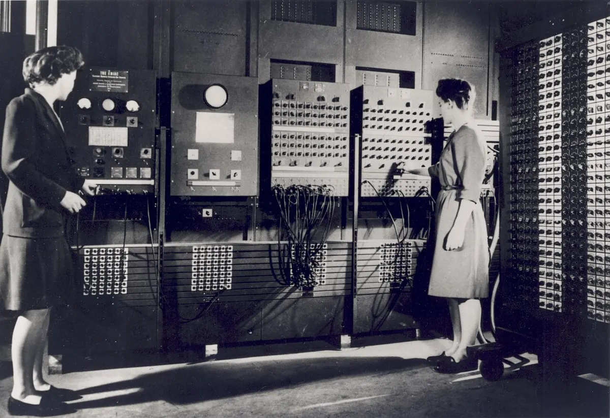 Two women setting switches on the huge control panels of an early computer that fills a room