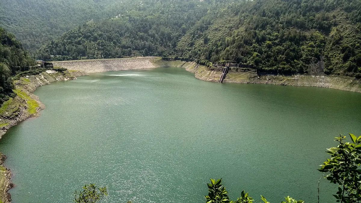 वनले ढाकिएका पहाडका बिचमा लामो हरियो ताल; तालको परतिर ढुङ्गाको बाँधले पानी रोकेको छ