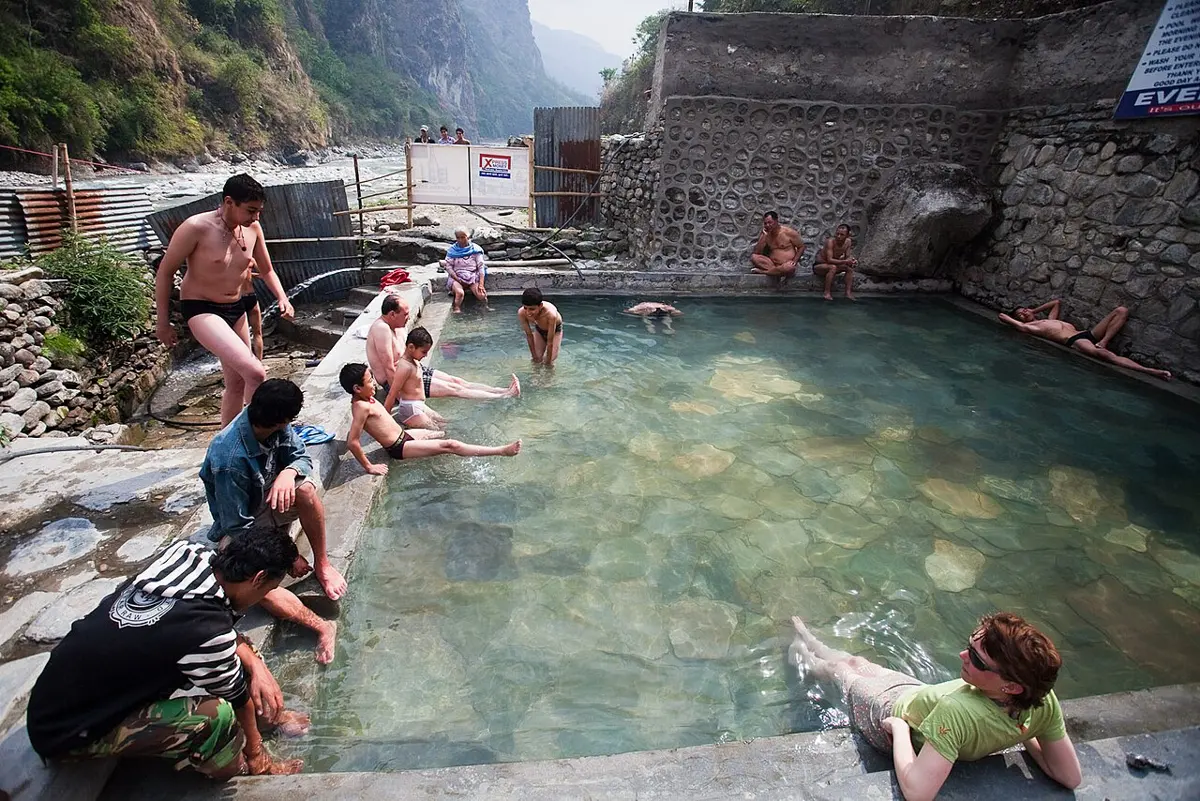 People sitting and bathing in a stone pool of naturally hot water beside a river in a steep valley