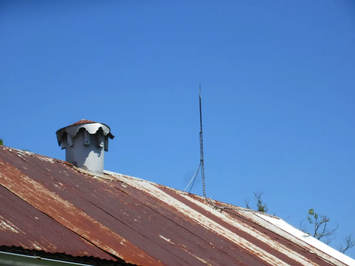 A thin, pointed metal rod standing up from the ridge of a rusty tin roof, with a cable running down from its base