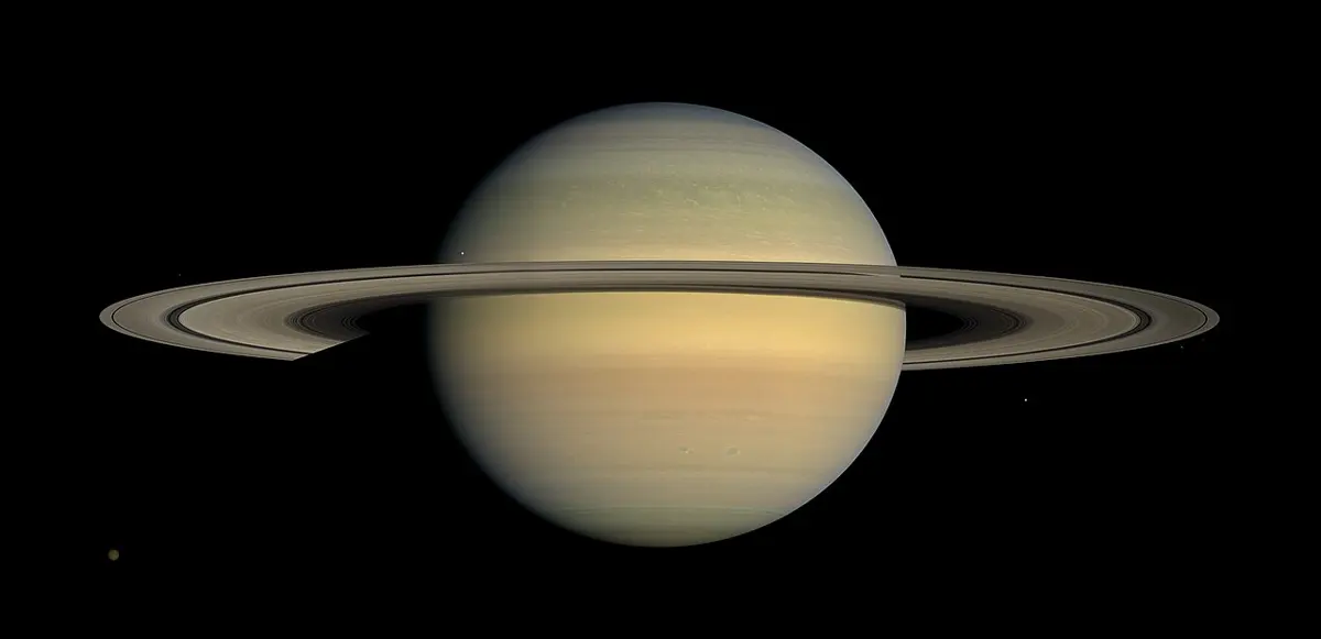 A pale golden planet with wide, thin rings seen almost edge-on, against black space