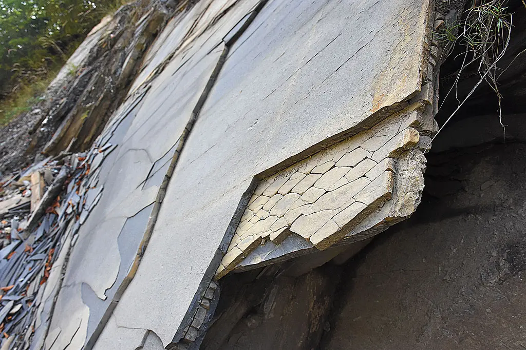 A gray rock face splitting into thin flat sheets, with broken flakes piled at its foot
