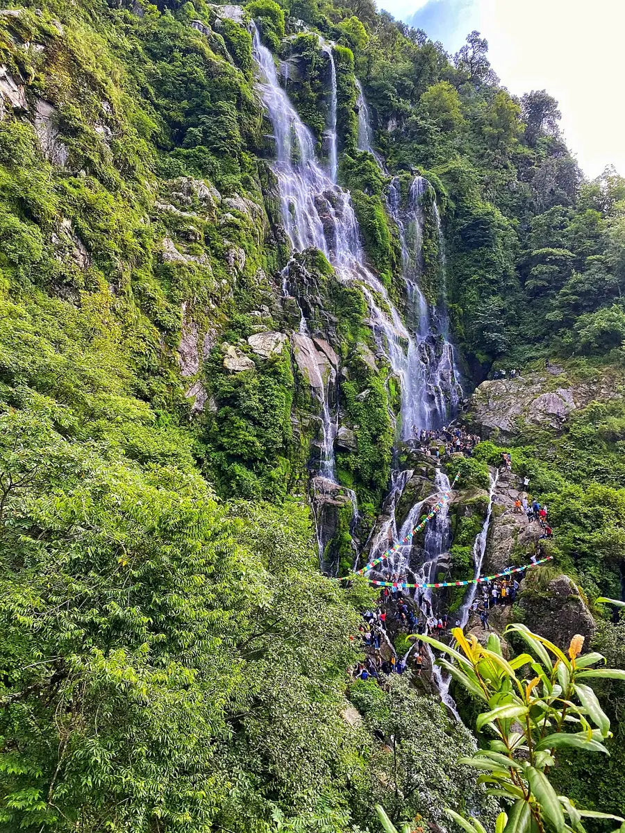A tall waterfall pouring in several white streams down a steep cliff covered in green forest, with tiny visitors at its foot