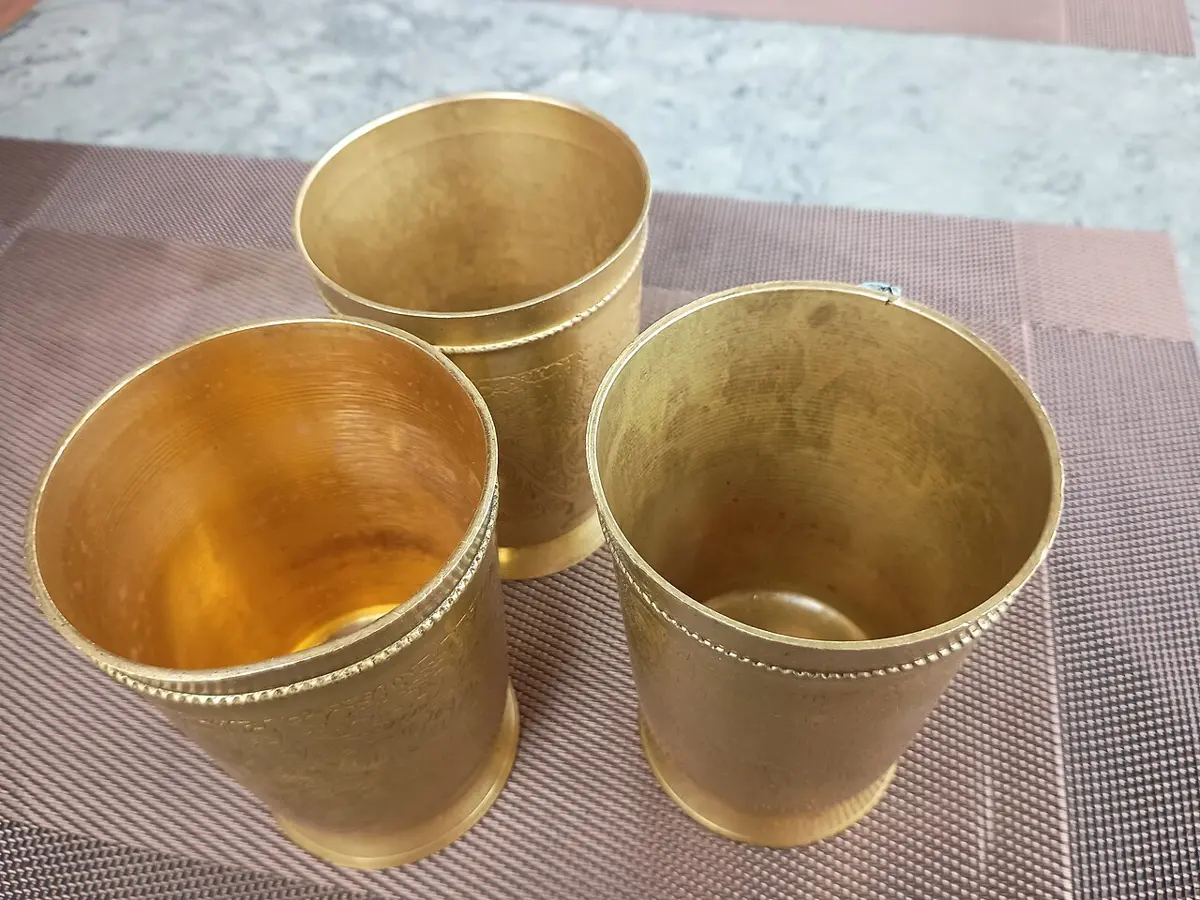 Three golden-colored metal drinking glasses on a woven mat