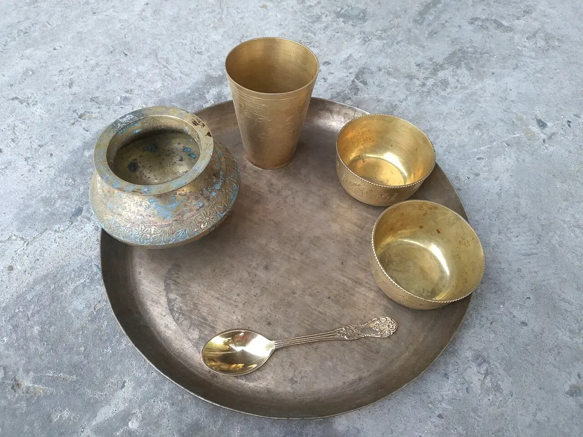 A large round brass plate with a brass glass, two bowls, a spoon and a small pot
