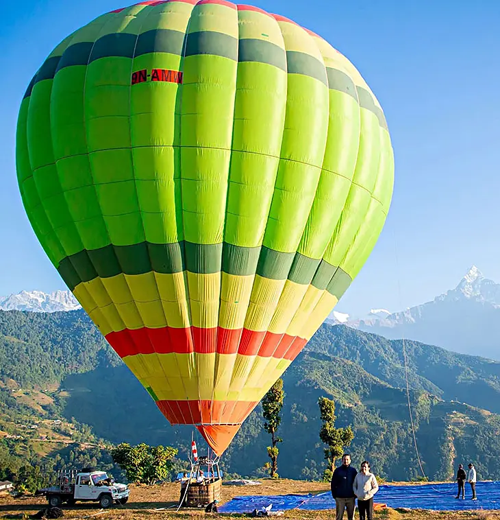 A big green and yellow hot air balloon, filled and ready to take off from a hilltop, with snowy mountains behind