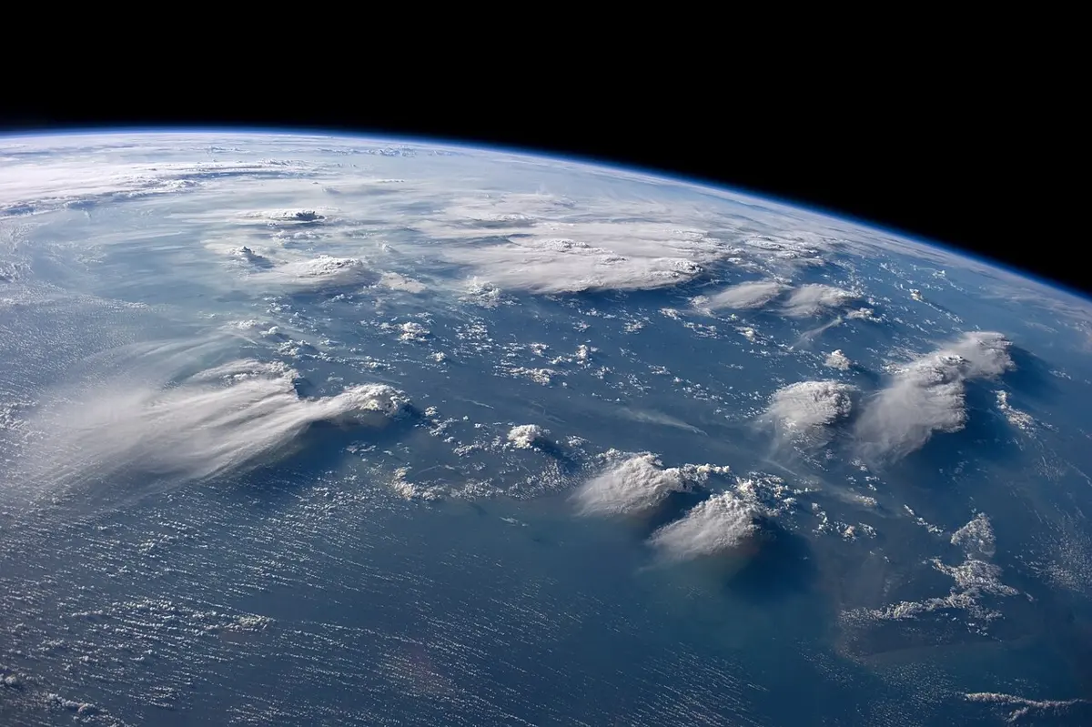 Seen from space, tall white thunderclouds tower over a band of lower clouds above a dark blue sea