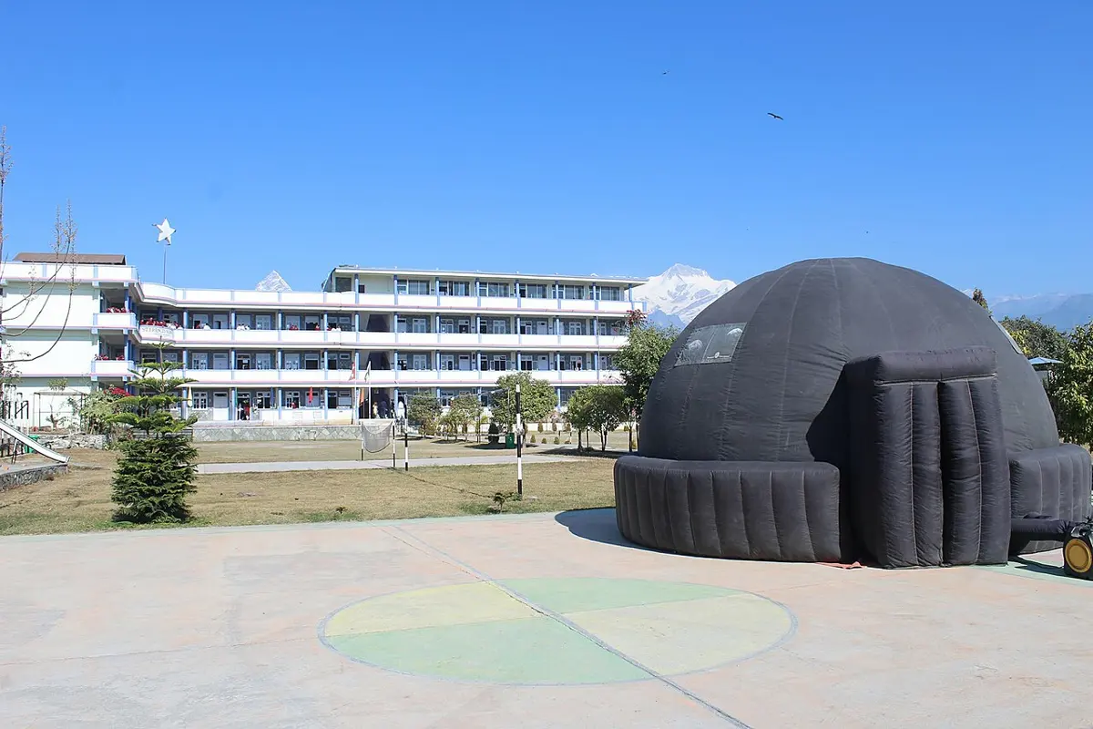 A large black inflatable dome with a tunnel entrance in a school courtyard, a white school building behind it and snowy mountains on the horizon under a clear blue sky