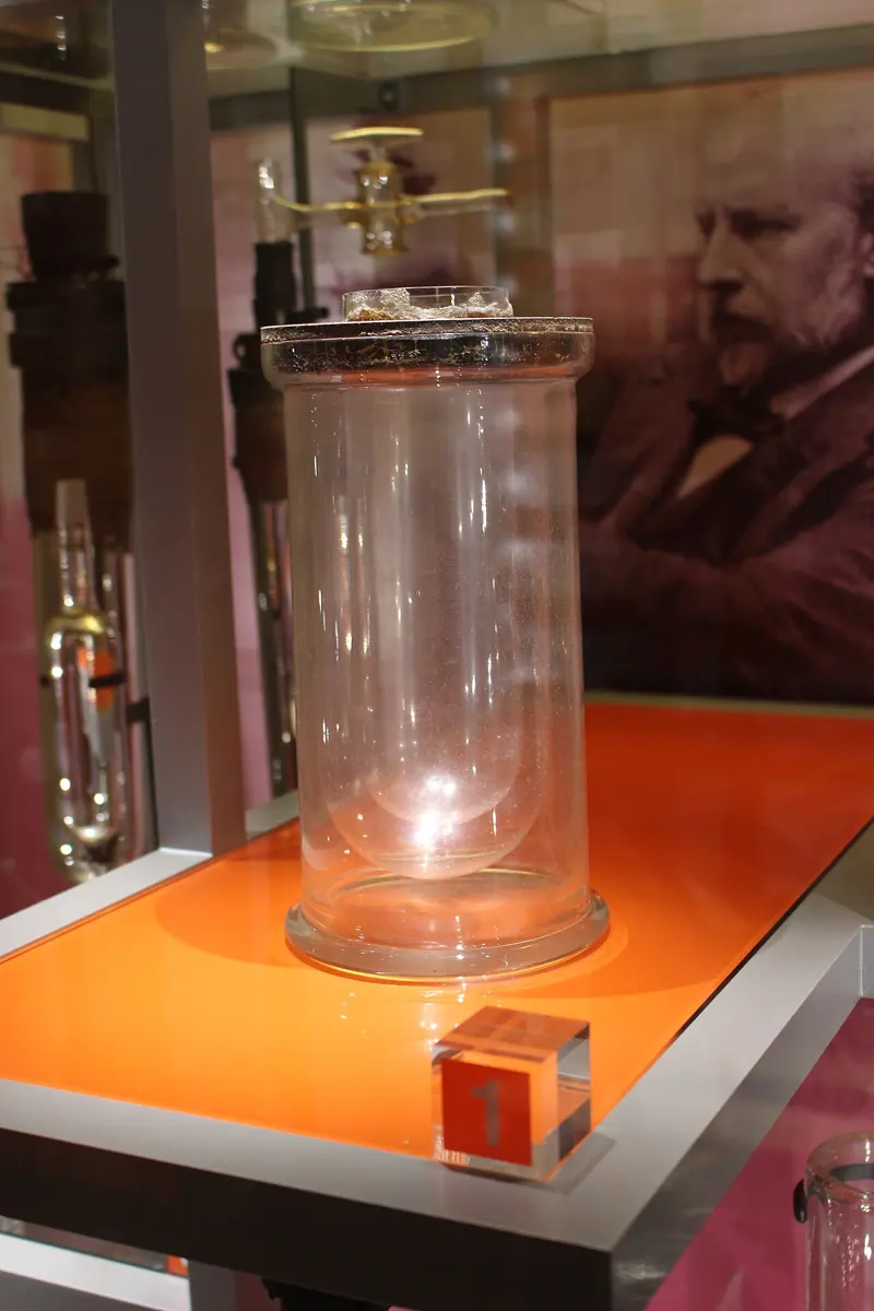 A tall glass vacuum flask in a museum case, with a painted portrait of a bearded man behind it
