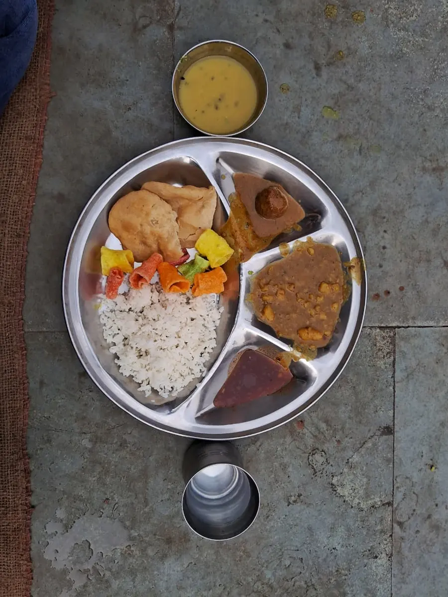 A meal of rice, dal, vegetables and roti served on a shiny round stainless-steel plate with compartments, with a steel bowl and glass