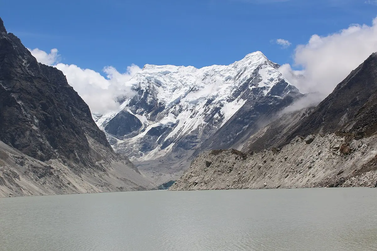 A long, gray-green lake in a bare, rocky Himalayan valley, below snowy mountains