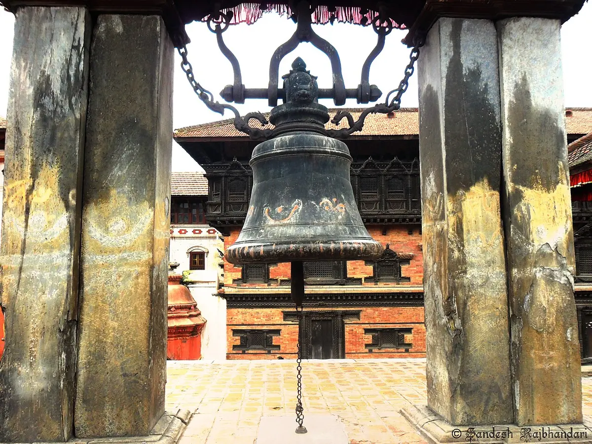 A large dark metal bell hanging from an iron frame between two thick stone pillars, with old brick buildings behind