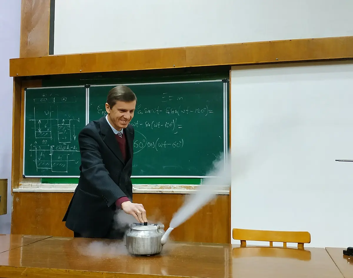 A teacher in a classroom with a metal kettle of liquid nitrogen, a white cloud streaming from its spout