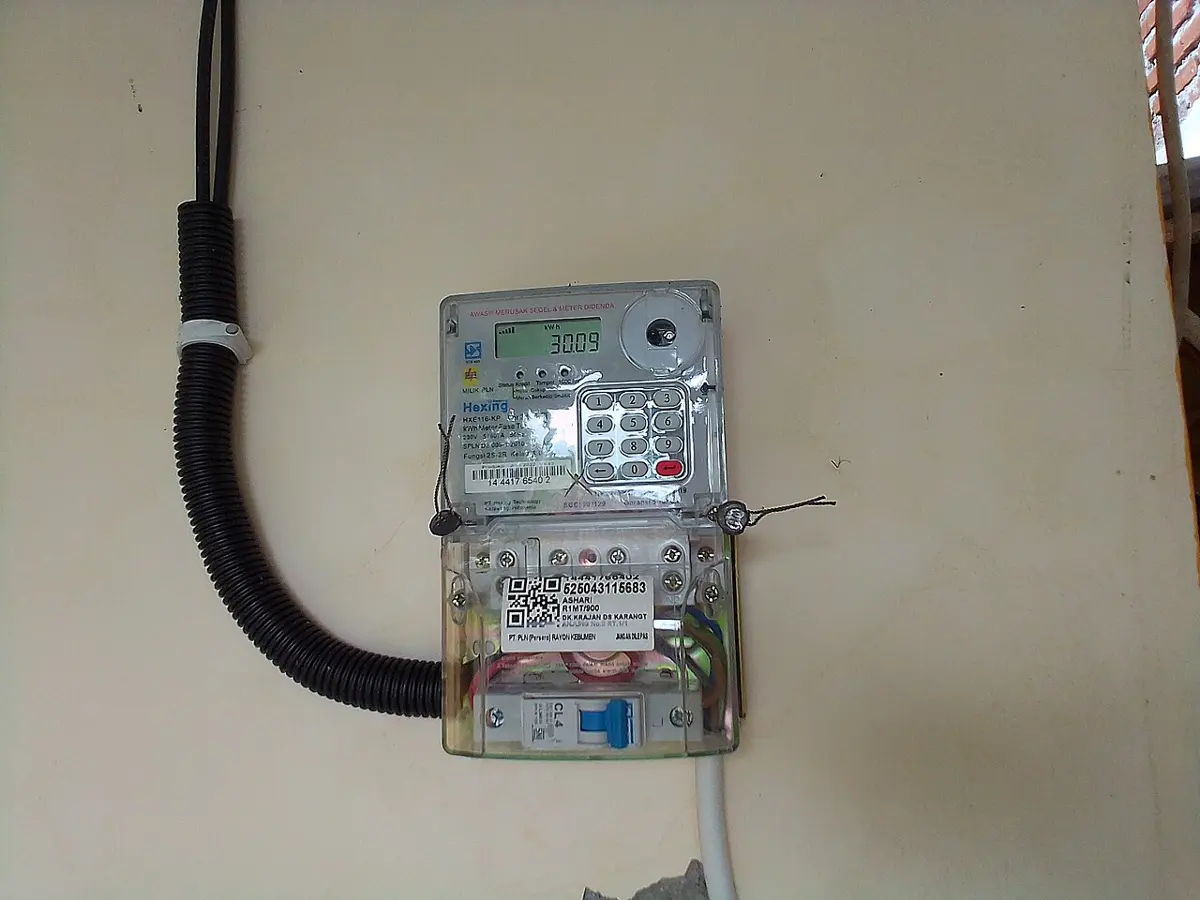 A digital electricity meter on a wall, with a small display reading in kWh, a keypad, and a small circuit breaker below it