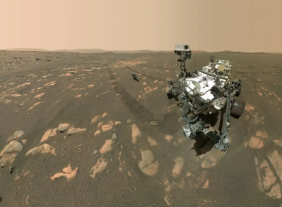 A six-wheeled robot vehicle on reddish rocky ground, with a small helicopter standing on the ground behind it