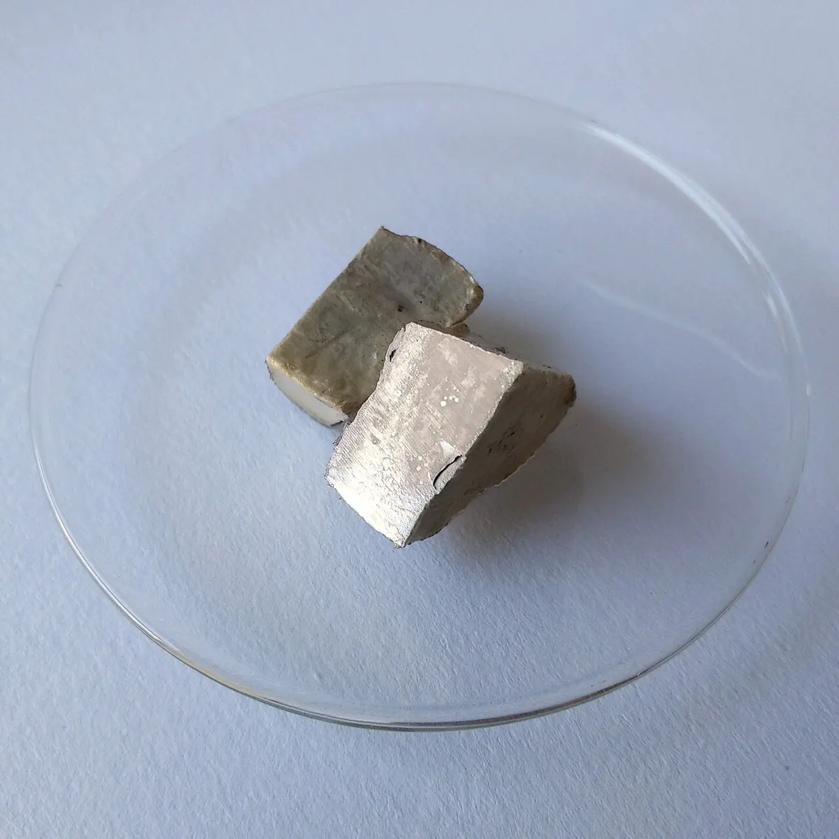A lump of sodium on a glass dish: one freshly cut face shines silvery, the rest is covered in a dull brownish crust