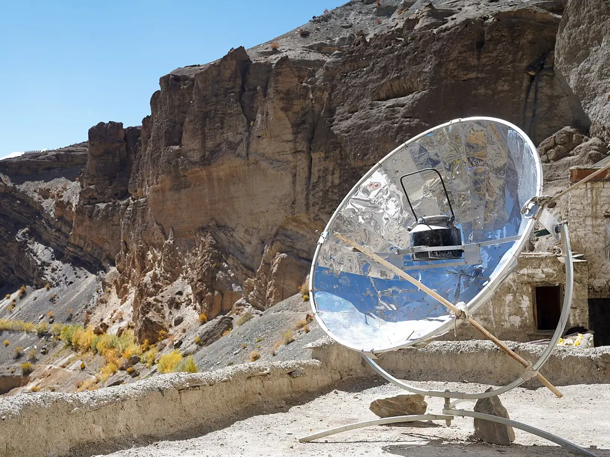 A large dish of shiny mirror panels on a stand, with a kettle held at its center, outside a Himalayan monastery