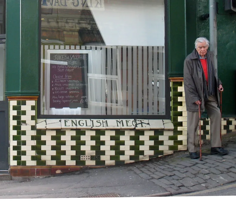 An old man with a walking stick standing by a café whose wall is tiled in rows of dark and light tiles