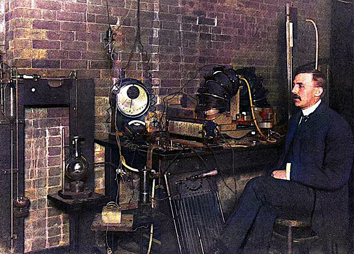 A man with a mustache in a dark suit sitting on a stool beside a laboratory bench crowded with electrical instruments, a large dial, glass flasks and wires, against a brick wall