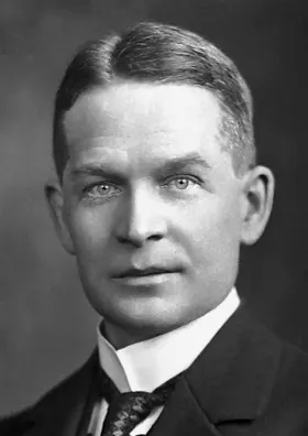 A black-and-white portrait of a man with short, neatly parted hair, in a dark jacket, white collar and tie