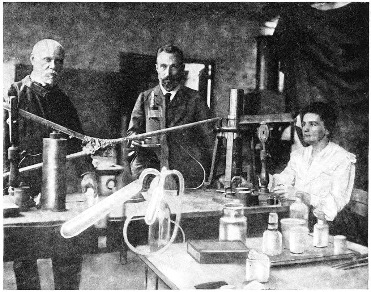 Marie Curie seated at a laboratory bench on the right, with Pierre Curie standing in the middle and an older bald man on the left, among glass tubes, bottles and a tall measuring instrument