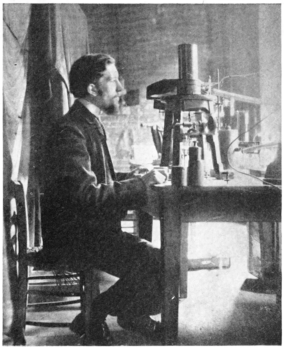 A bearded man in a dark jacket sitting at a laboratory table, beside a tall instrument on a stand with a metal cylinder on top, in a dim room