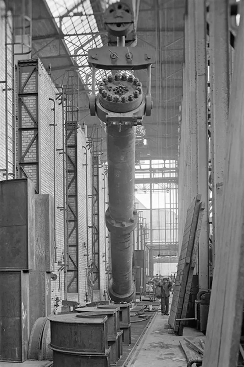 An old black-and-white photograph of a tall, thick metal tube, much taller than a person, hanging upright from a crane inside a large factory hall