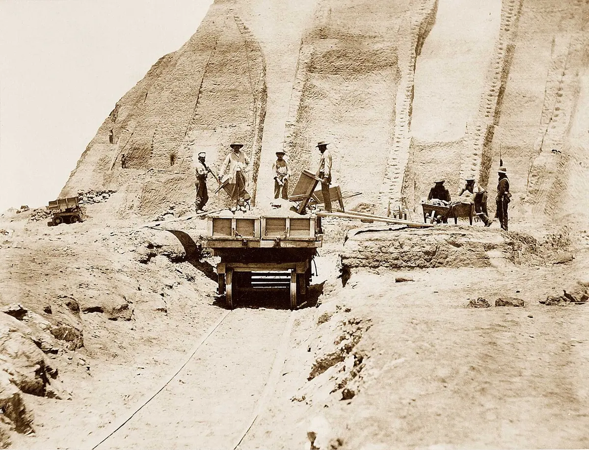 An old sepia photograph of workers with shovels and a cart on rails, digging into a huge pale cliff of guano on a bare island