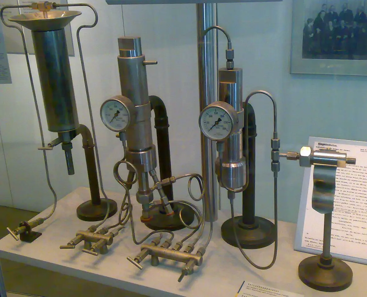 A museum display case holding laboratory apparatus: metal cylinders standing upright on stands, joined by thin metal pipes and valves, with two round pressure gauges