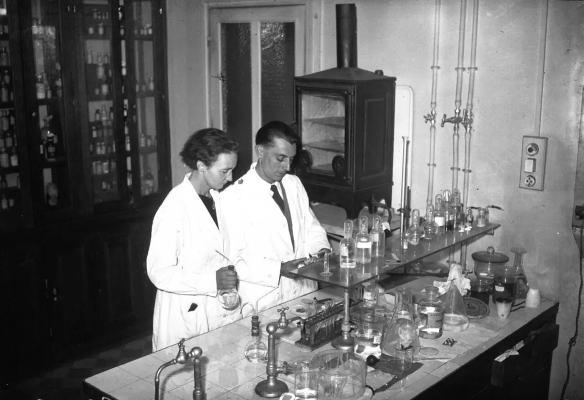 A woman and a man in white laboratory coats working at a tiled laboratory bench covered with glass bottles and flasks, with tall cupboards of bottles behind them