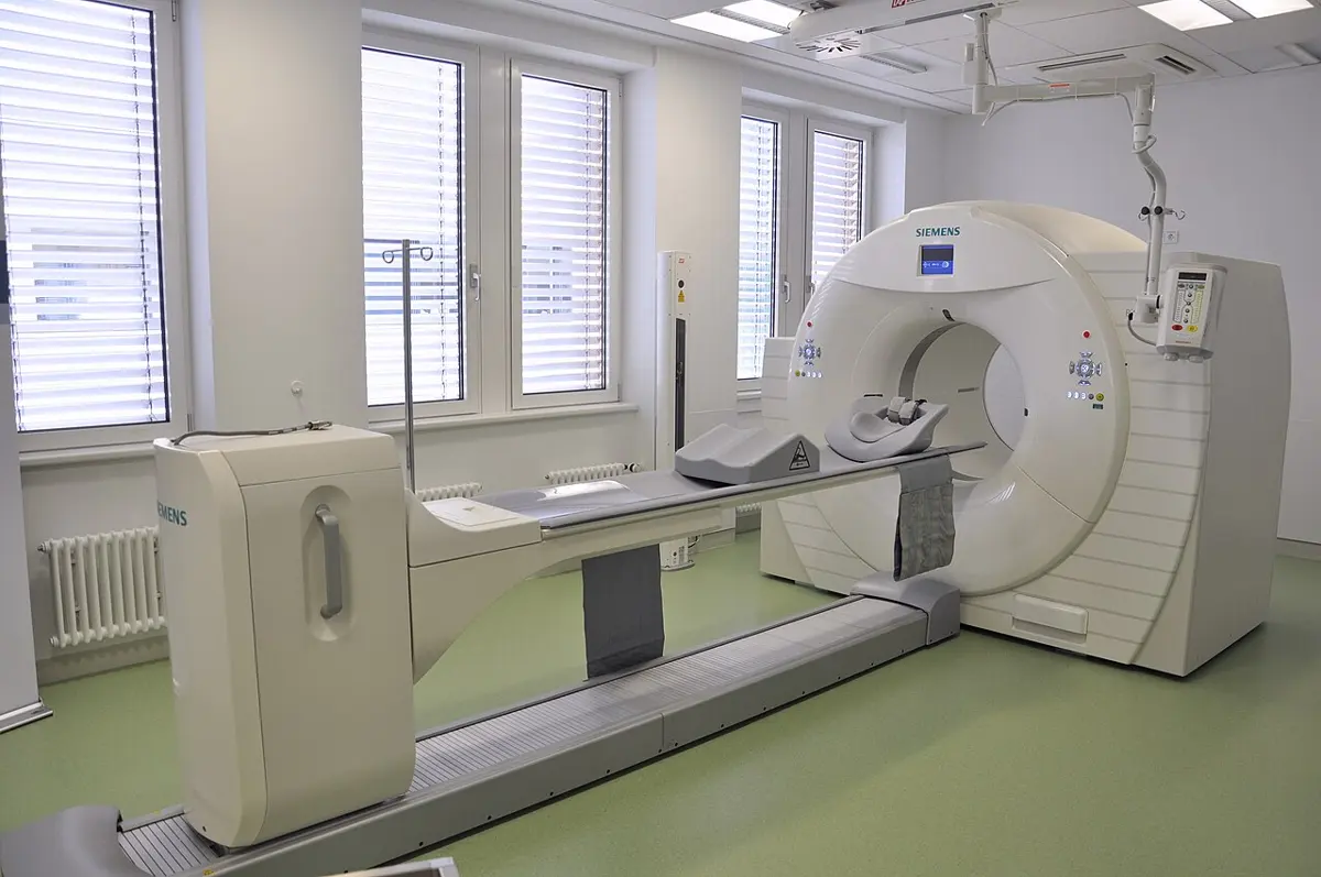 A large white ring-shaped scanner in a bright hospital room, with a narrow patient bed that slides into the hole in the middle