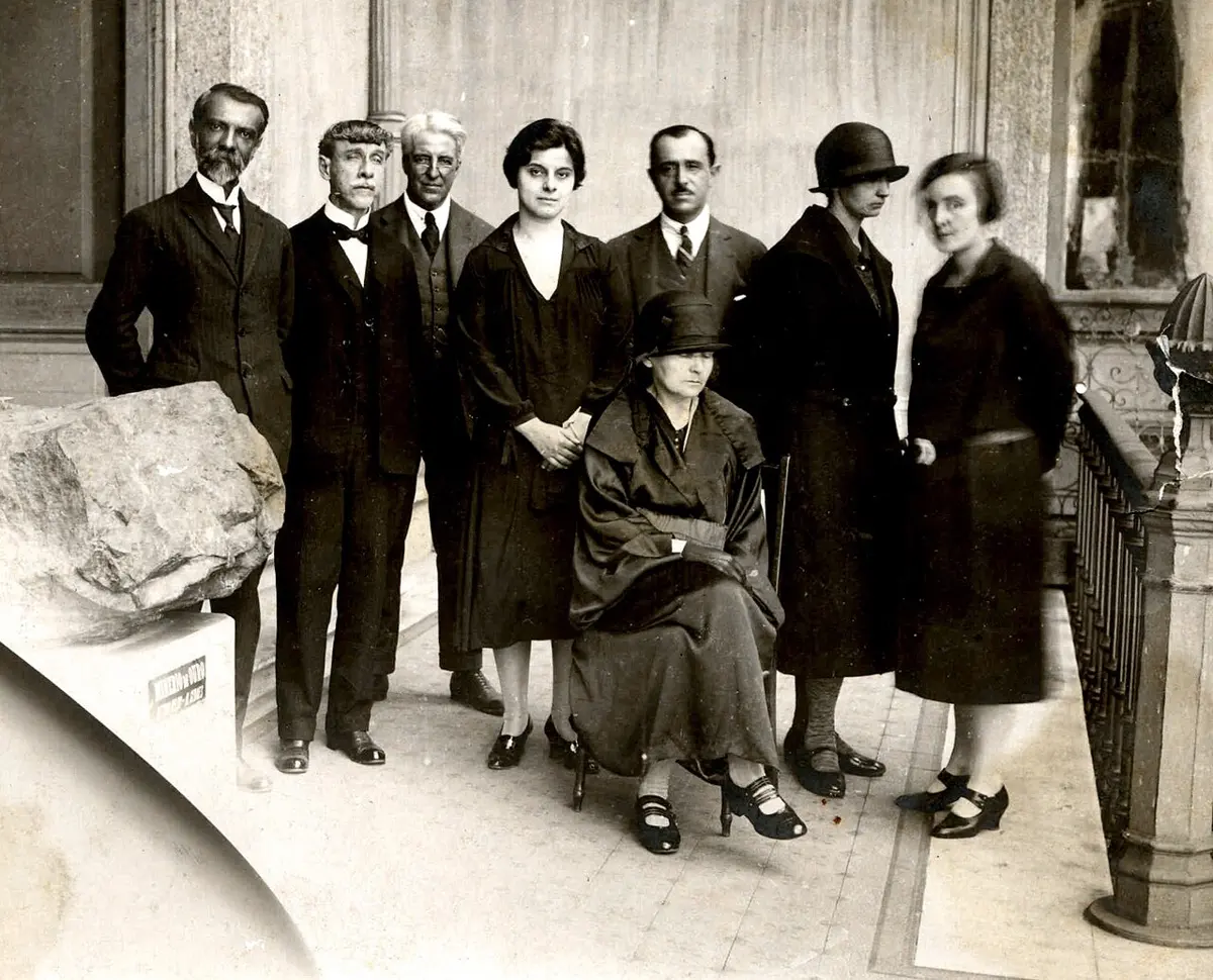 A group of eight people in dark 1920s clothes on a museum balcony: an older woman in a hat seated in the middle, with women and men in suits standing around her, beside a large rock on a stand