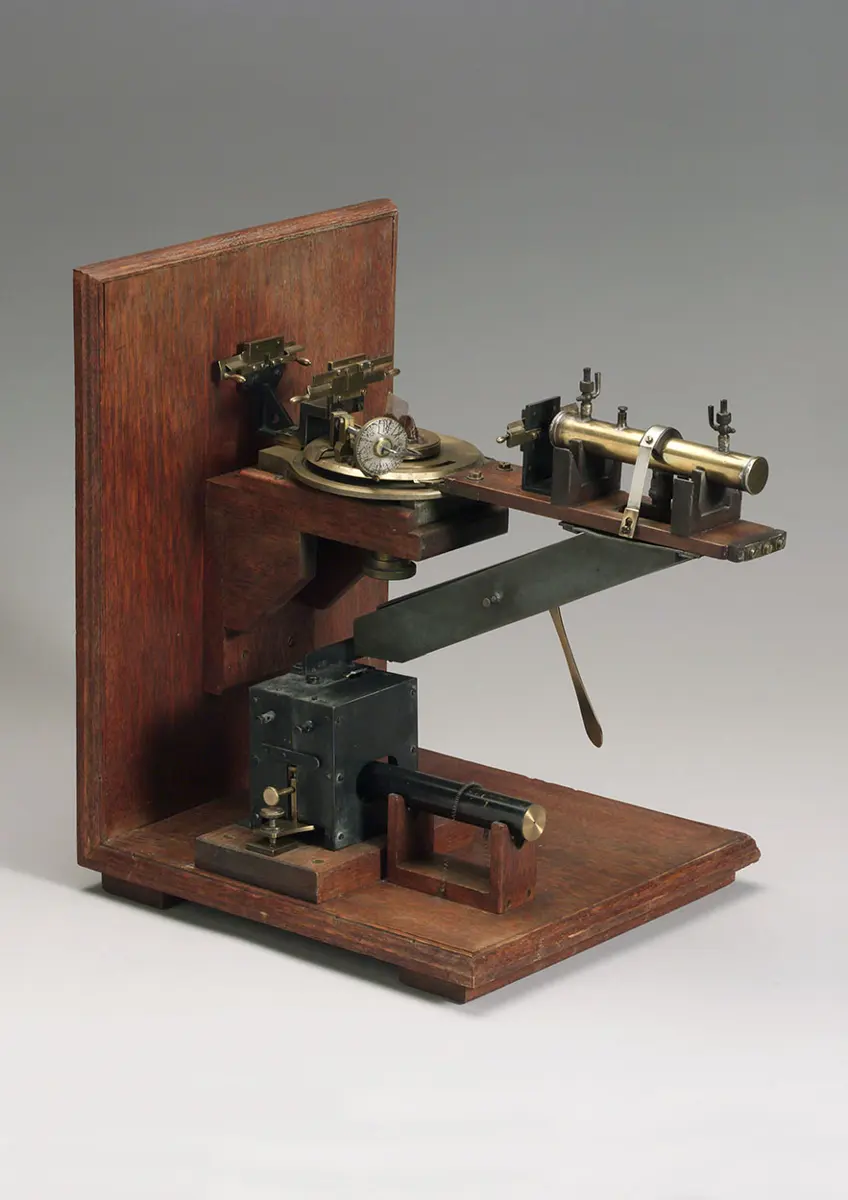 An old instrument of brass and dark metal on a wooden base, with a small table in the middle that can turn, a tube pointing at it and a long arm swinging round it
