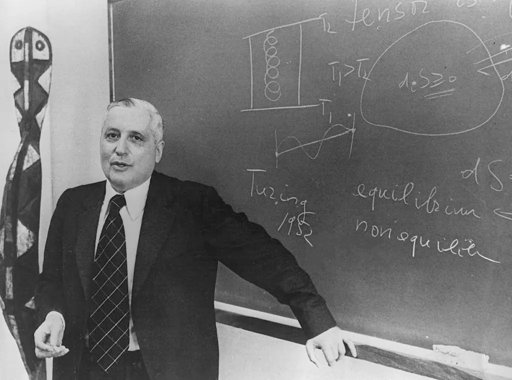 Black-and-white photo of a man in a dark suit and tie standing at a blackboard covered in chalk notes and diagrams