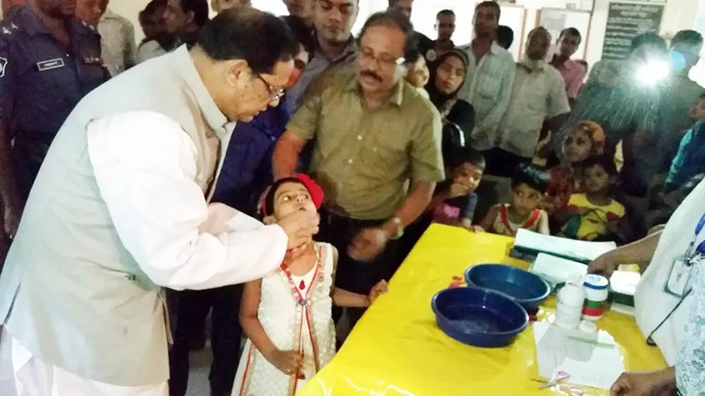 A man in a white kurta and gray waistcoat gives a vitamin A capsule to a small girl, who opens her mouth, while a crowd of adults and children watches
