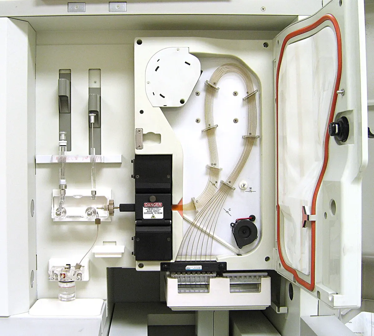 The open front of a cream-colored laboratory machine: a bundle of thin, hair-like tubes loops across a white panel into a black block with a laser danger sign, and glass syringes stand on the left