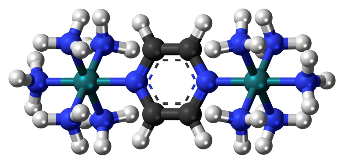 A ball-and-stick model on a black background: two green-blue metal atoms, each surrounded by blue and white groups, joined in the middle by a six-sided ring of black and blue atoms