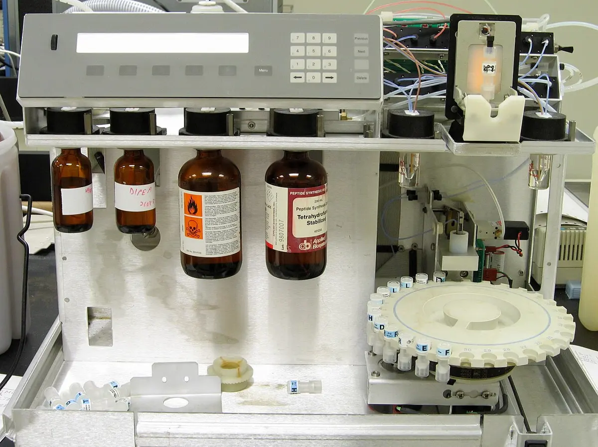 A laboratory machine with its front open: brown glass bottles of liquids hang in a row, tubes run to valves at the top, and on the right a round tray holds many small labeled vials