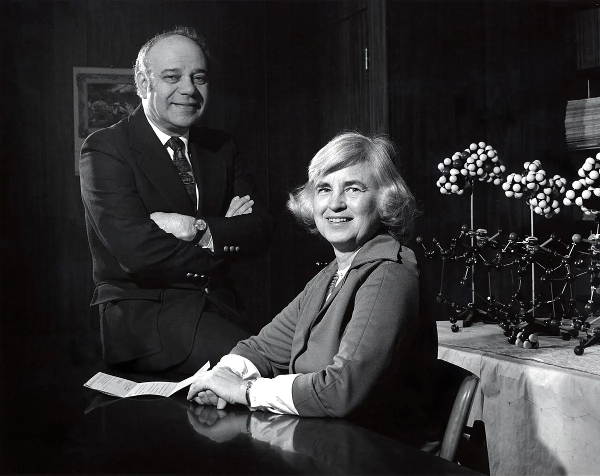 A black-and-white photo of a smiling man in a suit standing with folded arms behind a smiling woman seated at a table; beside them stand ball-and-stick models of molecules
