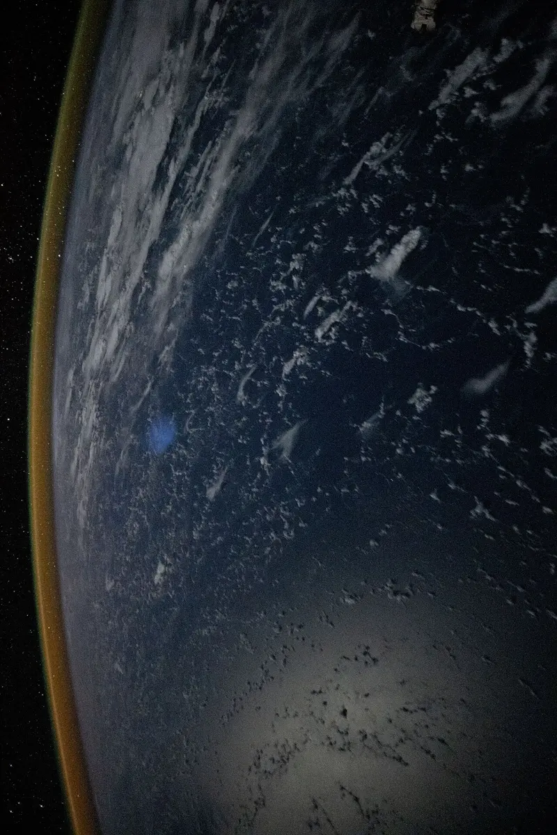 The curved edge of the Earth at night seen from space, clouds faintly lit below, with a thin orange-yellow band of glow lying along the horizon against the starry sky