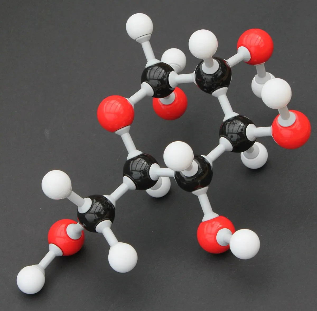 A ball-and-stick model of a glucose molecule on a dark background: black carbon balls and one red oxygen ball joined in a ring, with more red oxygen and white hydrogen balls sticking out